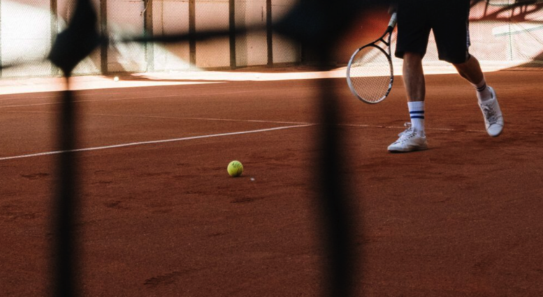 A tennis player in shorts and white shoes stands on a clay court near a tennis ball, illustrating how descriptive trademarks can be as straightforward as the visual elements used to identify and define a brand's trademark definition.