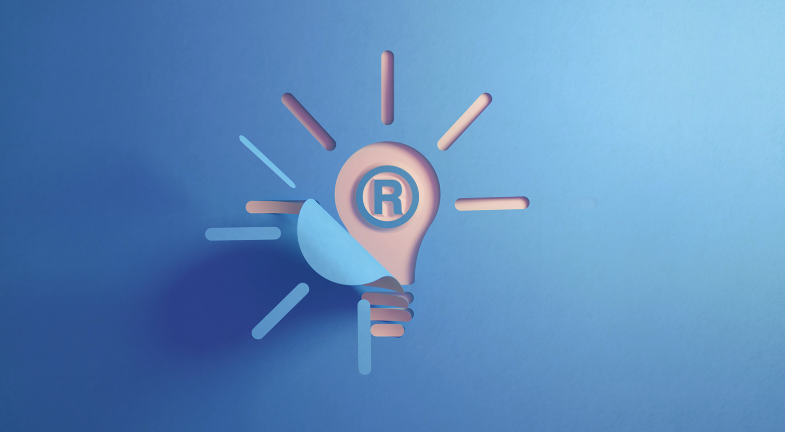 A stylized pink lightbulb with a registered trademark symbol illustrates the concept of a descriptive trademark on a blue background, with radiating lines representing light.