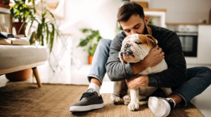 A man sits on a rug in a living room, hugging his English bulldog, the loyal companion who comforts him as he reflects on the thought of dying without heirs.