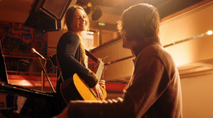 A woman with a guitar smiles at a man playing piano on a warmly lit stage, performing "Dying Without Heirs," as microphones and an audience area set the scene in the background.