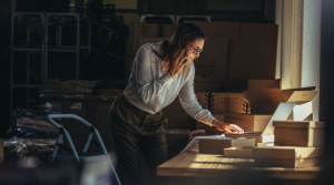 A woman stands in a warehouse packing area, talking on the phone and using a laptop, perhaps discussing an urgent order about inventory from someone dying without heirs, surrounded by cardboard boxes and packaging materials.
