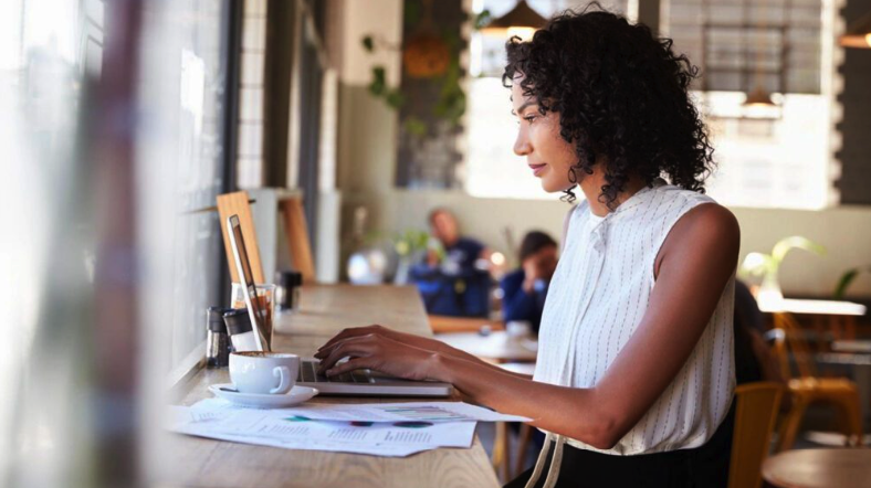 A woman sits at a counter in a cafe, working on a laptop with documents about inheritance law and a cup of coffee in front of her.