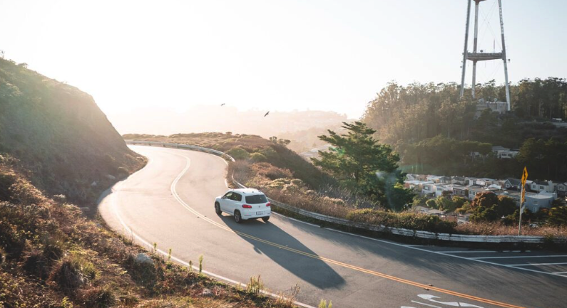A white car drives along a winding mountain road at sunrise, passing trees, houses, and a tall transmission tower—an evocative reminder of lonely journeys and legacies left behind, like dying without heirs.