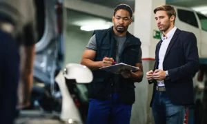 Two men stand by an open car hood in a garage; one holds a clipboard and pen, while the other listens. A vehicle is visible on a lift in the background.