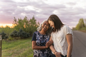 Two women stand arm-in-arm on a rural road at sunset, laughing together. Trees and greenery line the roadside, while a peaceful moment unfolds far from worries about auto insurance claims and damages.