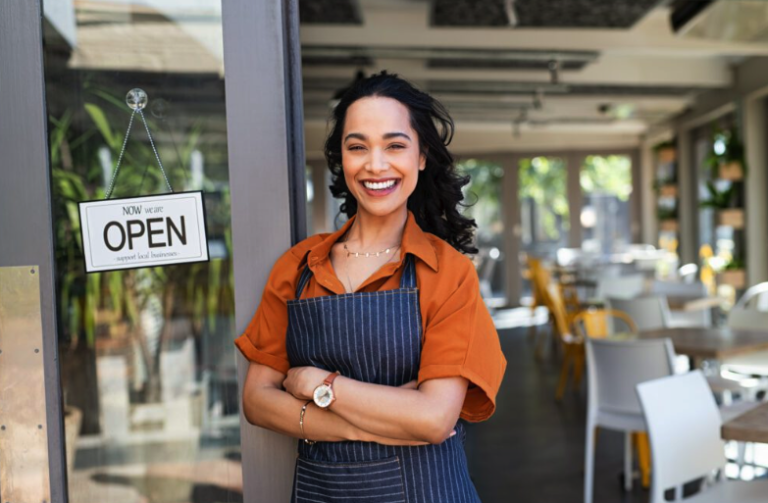 Woman wearing an apron stands smiling with arms crossed at the entrance of a café with an "open" sign, confidently ready to welcome guests—much like being prepared with third-party auto insurance for any unexpected events. Indoor seating and natural light are visible inside.