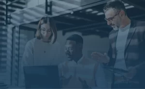 Three people in business attire are gathered around a laptop, discussing work in a modern office setting with natural light and industrial decor.