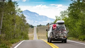 A van with two bicycles mounted on the back drives down a scenic, empty road surrounded by trees, with mountains in the background—an ideal scene for summer driving and a reminder to prioritize driver safety during the 100 Deadliest Days.