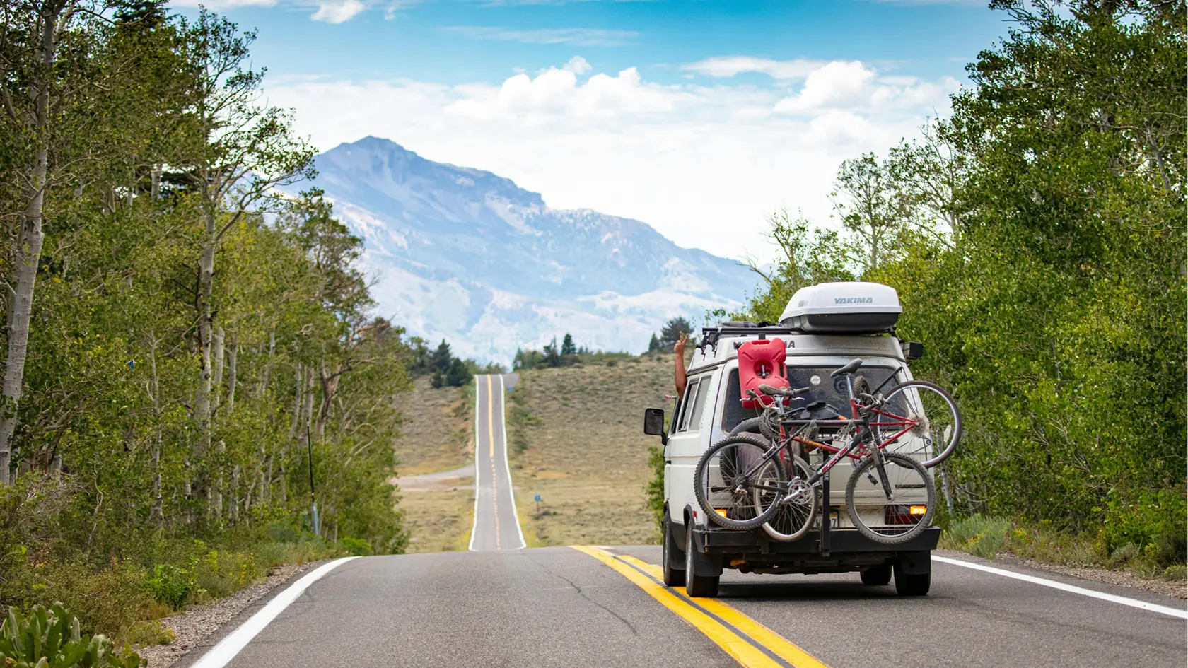 A van with two bicycles mounted on the back drives down a scenic, empty road surrounded by trees, with mountains in the background—an ideal scene for summer driving and a reminder to prioritize driver safety during the 100 Deadliest Days.
