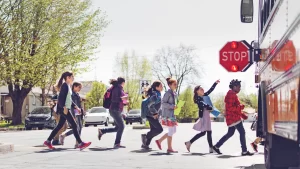 A group of schoolchildren cross the street toward a school bus with its stop sign extended on a sunny day, reminding every Texas driver about safe back to school driving during National Traffic Awareness Month.