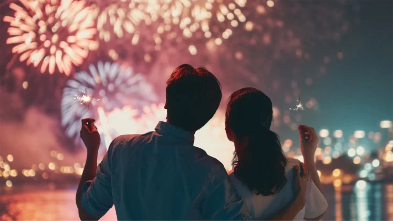 A couple stands by the water at night, holding sparklers and watching a large fireworks display over the city—Celebrate Safely with 4th of July safety tips for a memorable and secure evening.