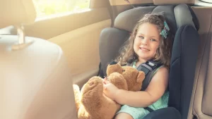 Young girl sitting in a car seat in the back of a vehicle, smiling and holding a brown teddy bear, illustrating child passenger safety and the importance of following Texas car seat laws.