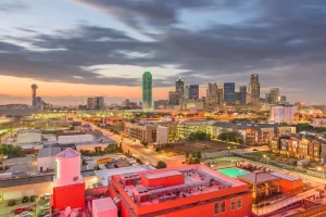 Dallas skyline at sunset with illuminated buildings, including the green-lit Reunion Tower, local families in residential areas below, and a cloudy sky overhead capture the vibrant spirit of Dallas evenings.