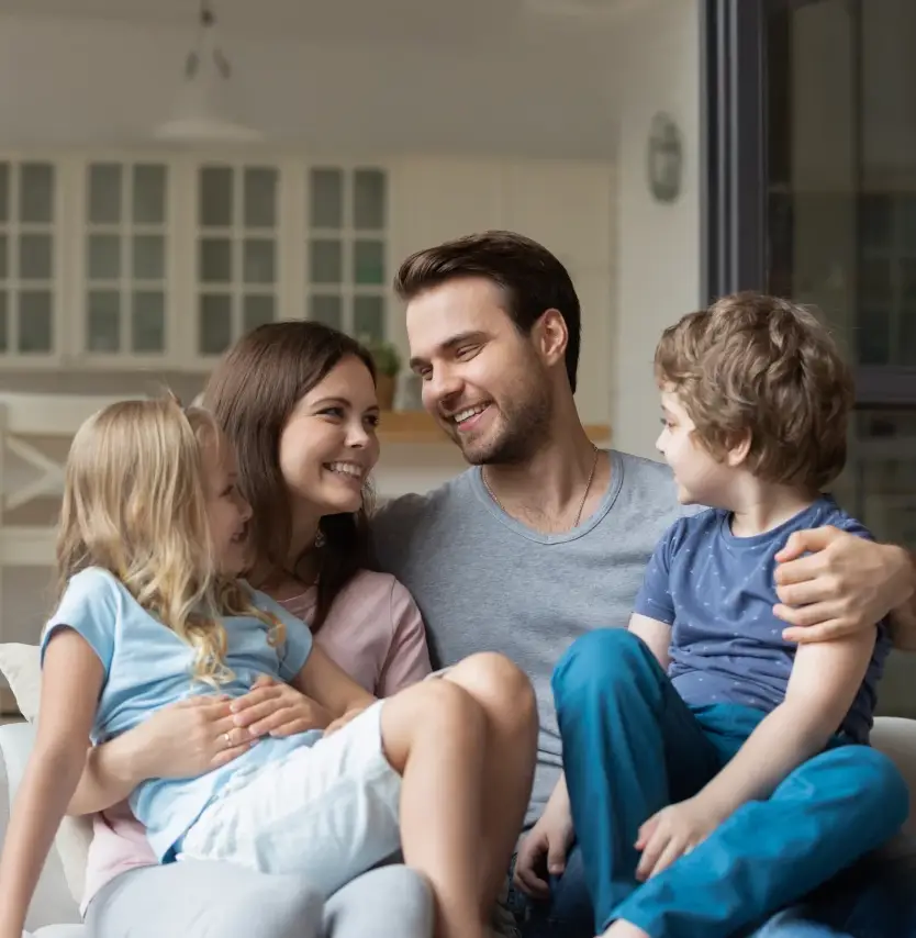 A man and woman sit closely together on a couch, smiling at each other, with a young girl and boy on their laps—all appearing happy and relaxed in a bright living room, enjoying the peace of mind that comes from thoughtful estate planning.