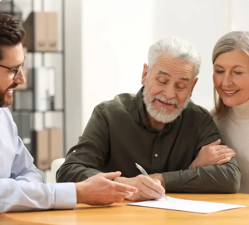 An older man signs an estate planning document at a table while a woman looks on, smiling, and a man in glasses points to the paper.