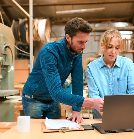 Two people in a woodworking shop look at a laptop together at a workbench, reviewing papers and a clipboard—perhaps discussing Business Law as it relates to their craft, with a mug nearby.