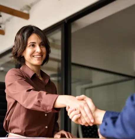 A woman in a brown shirt smiles while shaking hands with another person in an office setting, reflecting a positive outcome from a Business Law consultation.