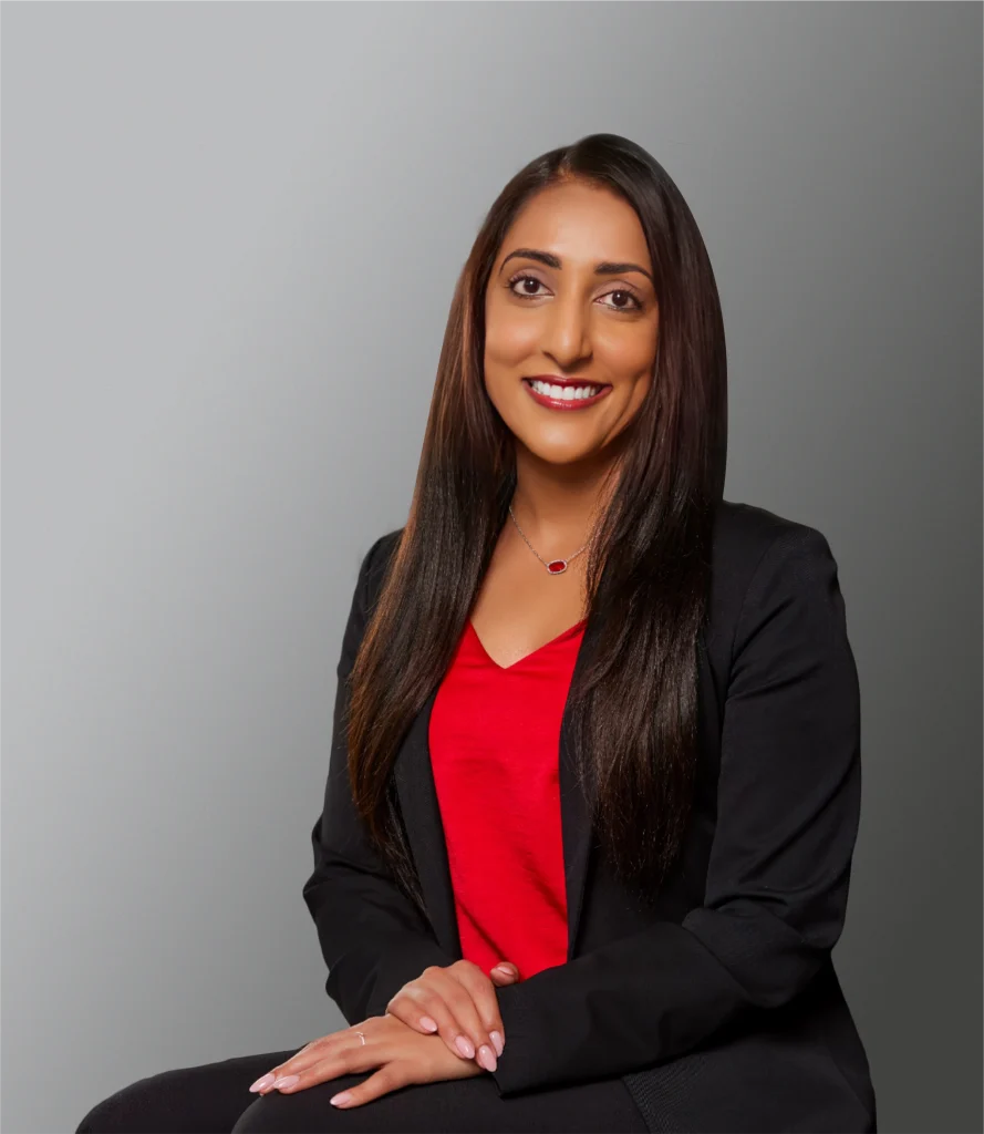 A woman with long straight hair, wearing a black blazer and red top, sits with hands folded and smiles at the camera against a plain gray background, exuding confidence perfect for an estate planning professional.