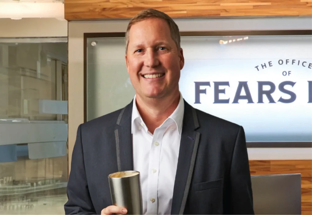 A man in a suit, holding a metal tumbler, smiles at the camera while standing before a sign that reads "The Office of Fears," perhaps contemplating the importance of estate planning.