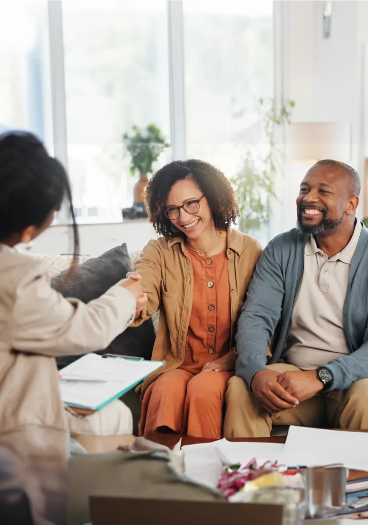 Two people sit on a couch, smiling, as one of them shakes hands with a person across the table during an estate planning meeting in a well-lit living room.